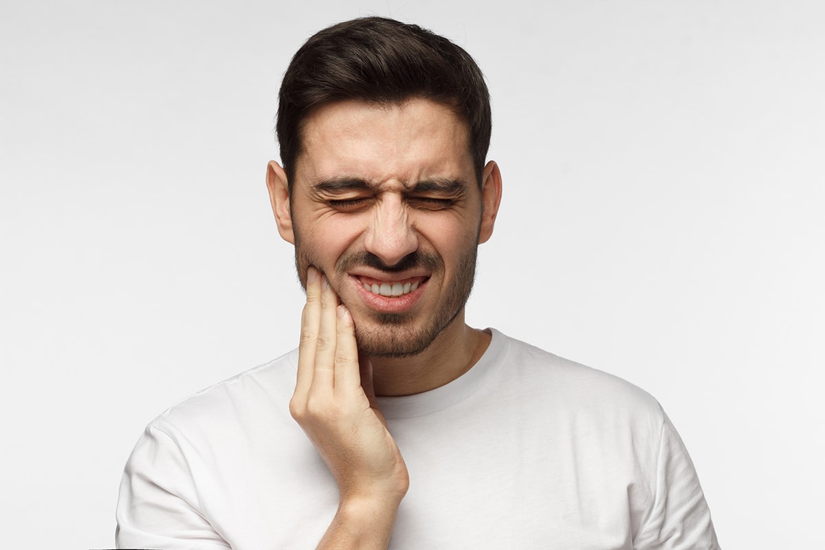 Closeup of young man isolated on gray background touching his face and closing eyes with expression of horrible suffer from health problem and aching tooth, showing dissatisfaction