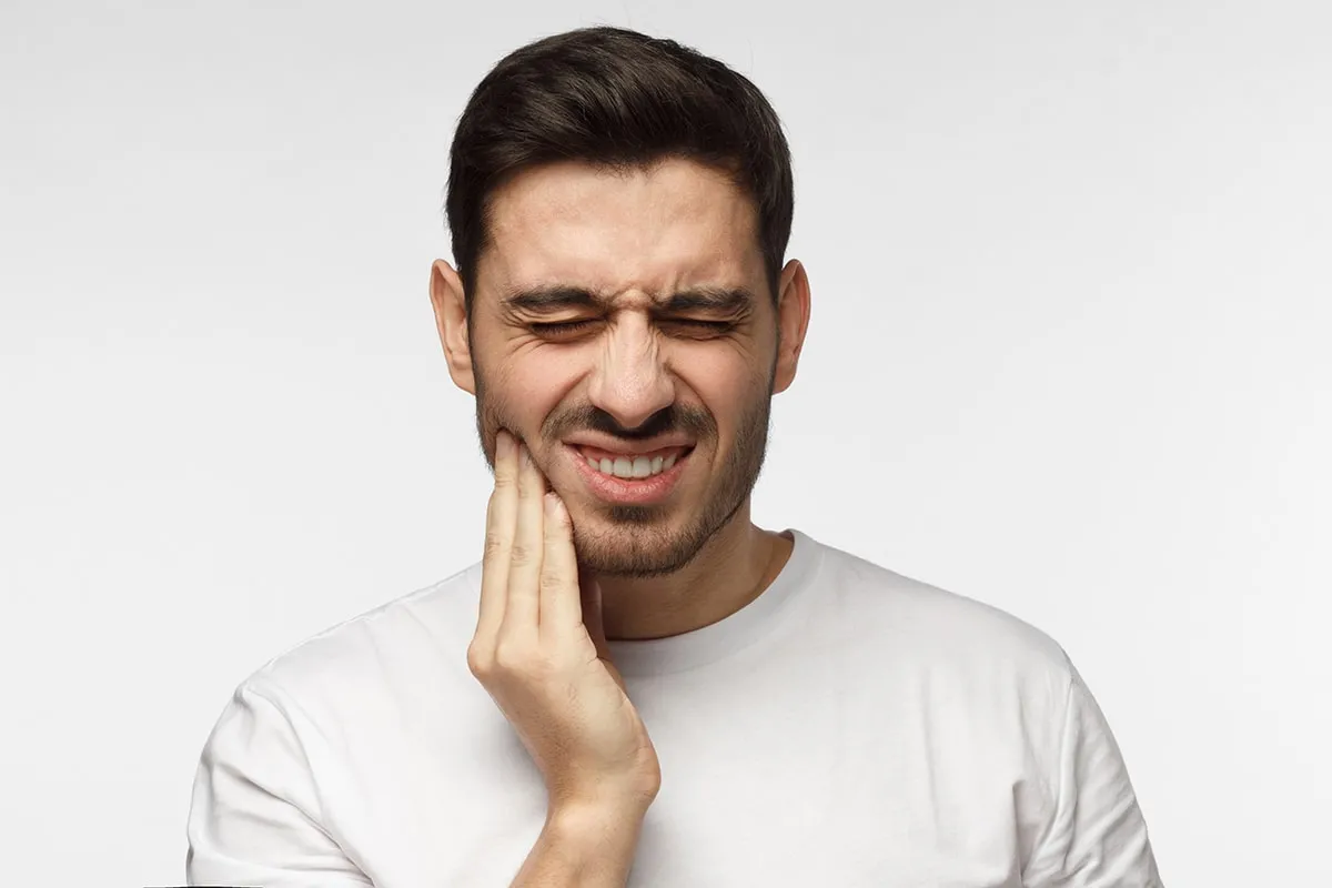 Closeup of young man isolated on gray background touching his face and closing eyes with expression of horrible suffer from health problem and aching tooth, showing dissatisfaction